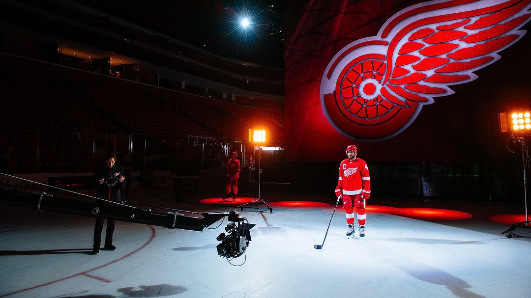 A lone Red Wings player lit on dark ice beneath a giant illuminated logo