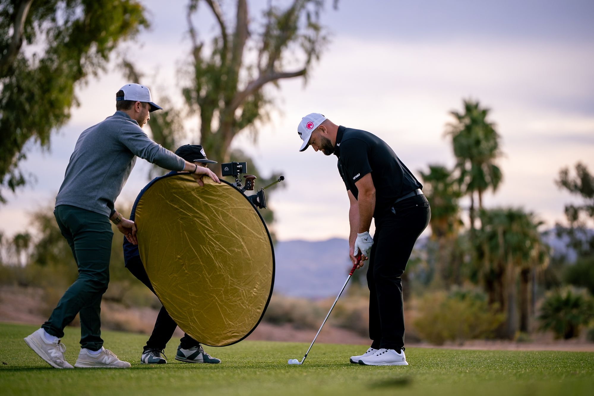 A crew member holds a gold reflector beside a golfer mid-swing on a desert course