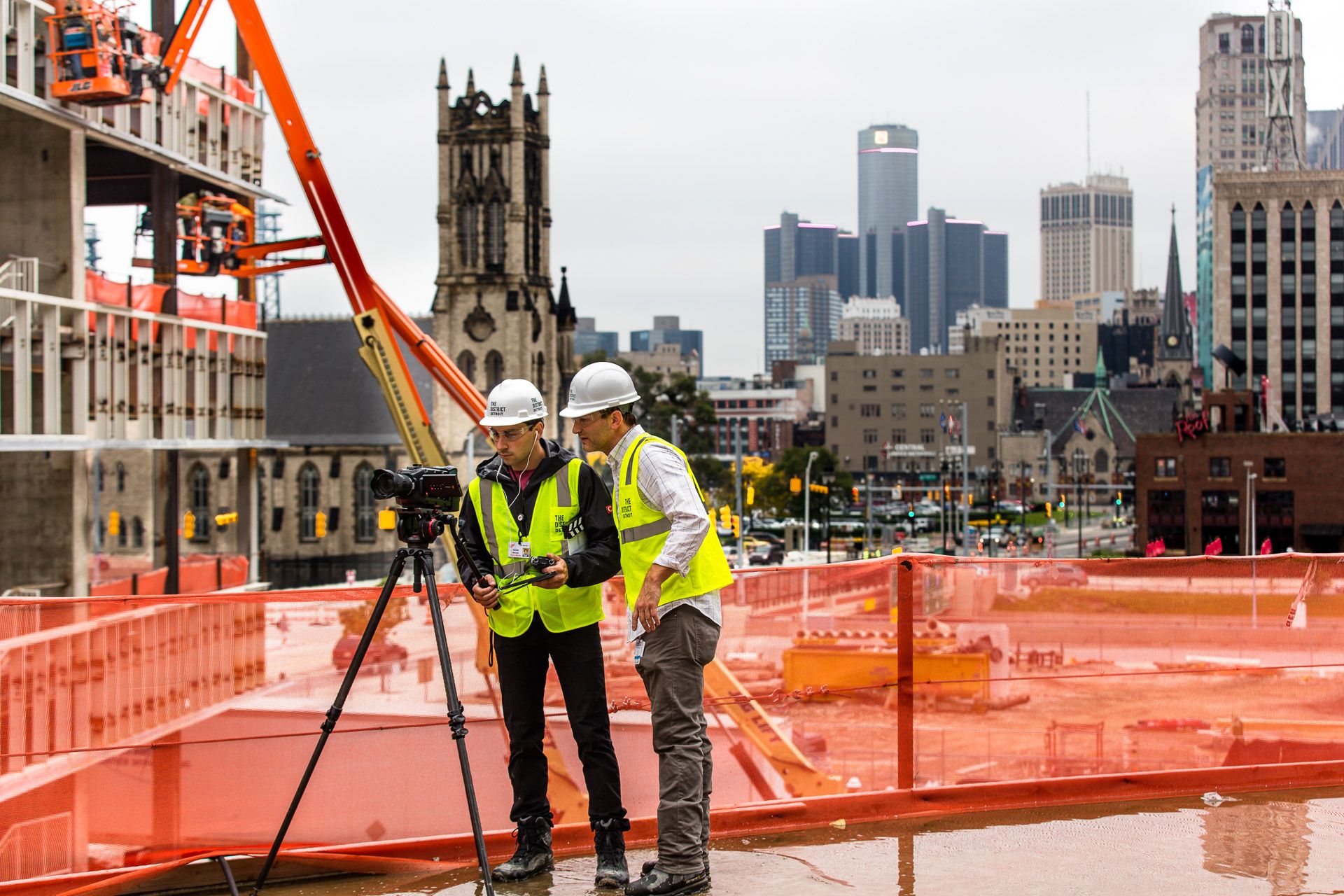 Two crew members in safety vests filming the Detroit skyline from the arena construction site
