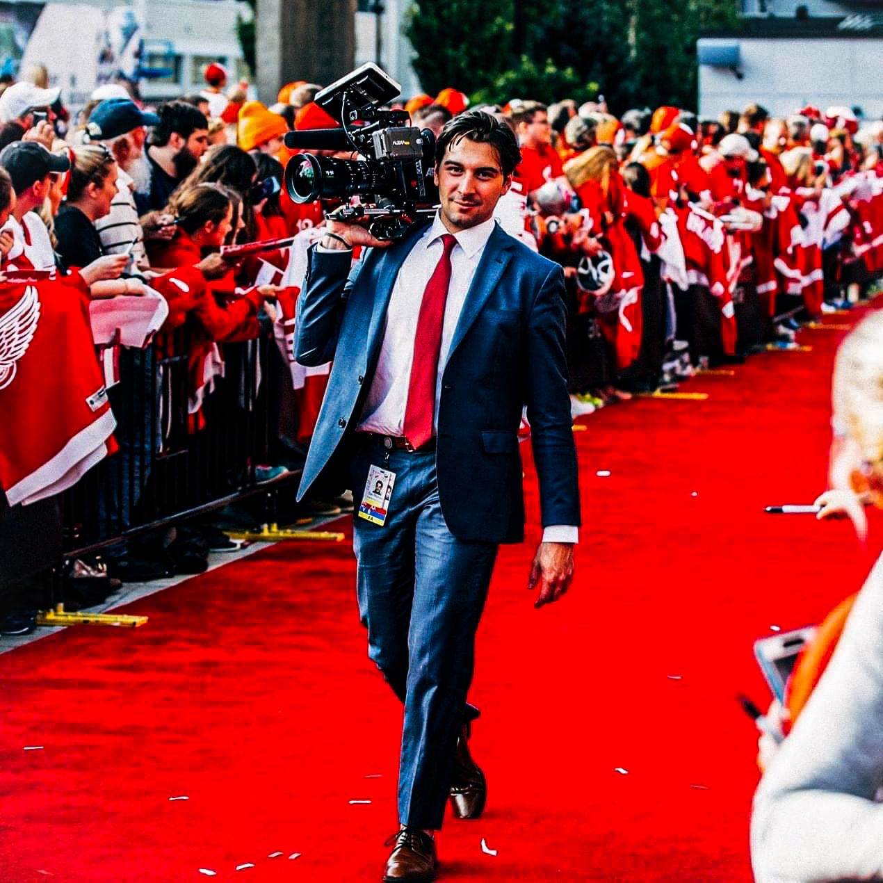 Dan in a suit walking the red carpet with a camera rig as fans line the barricades